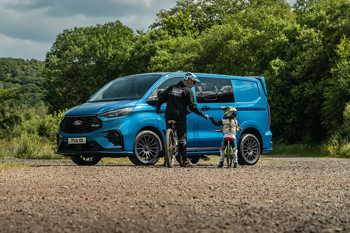 a blue ms-rt ford transit custom with a adult and child on bikes in front of it